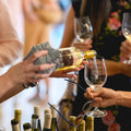 White wine being poured into glass at wine tasting event with multiple bottles and guests holding wine glasses